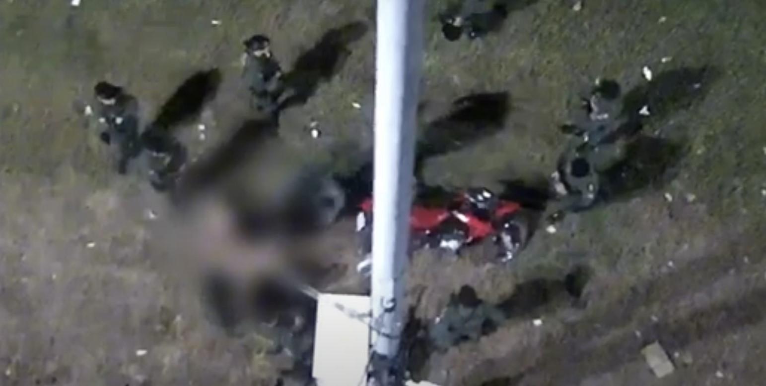 Imagem aérea de uma cena com várias pessoas e uma motocicleta ao redor de uma estrutura de lâmpada, à noite.