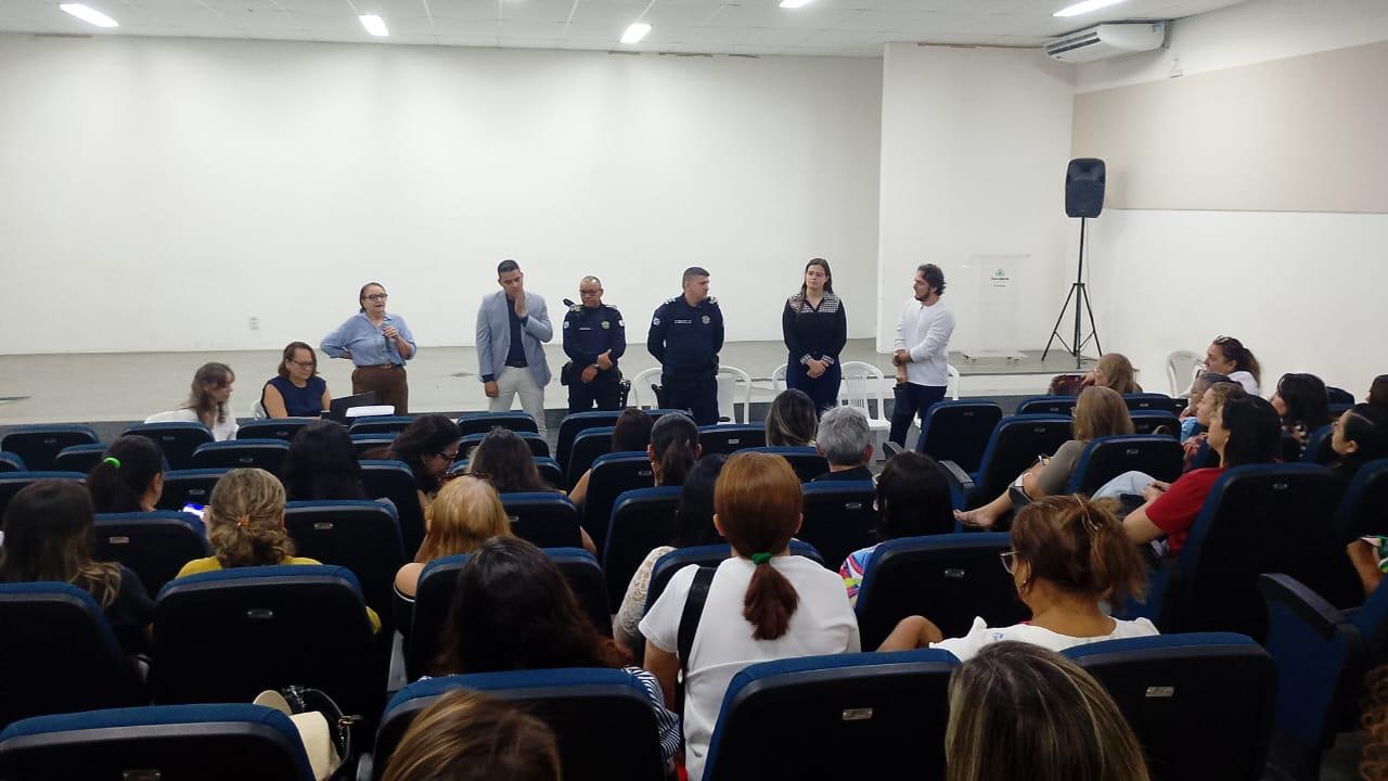 Pessoas sentadas em um auditório observam seis pessoas em pé à frente, participando de uma fala ou apresentação. Entre elas, há duas pessoas vestidas com uniforme de guarda municipal e quatro em roupas sociais. O ambiente é claro, com cadeiras azuis ocupadas pelo público.
