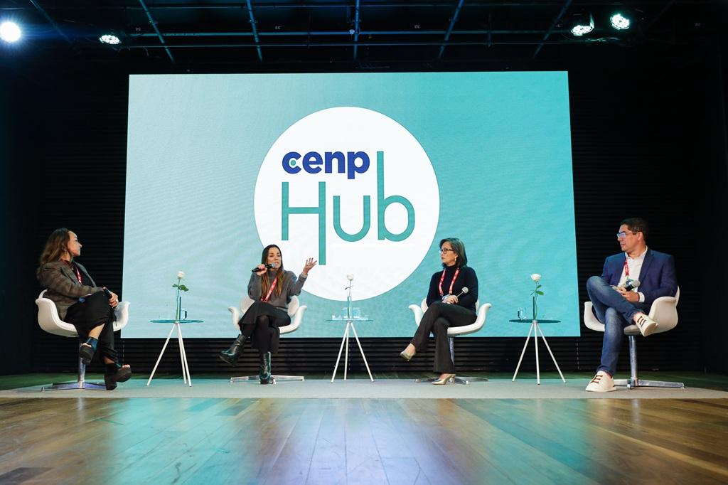 Quatro pessoas participam de um painel no evento CenpHub, sentadas em cadeiras brancas no palco. Atrás delas, um telão exibe o logotipo “cenp Hub” em destaque.