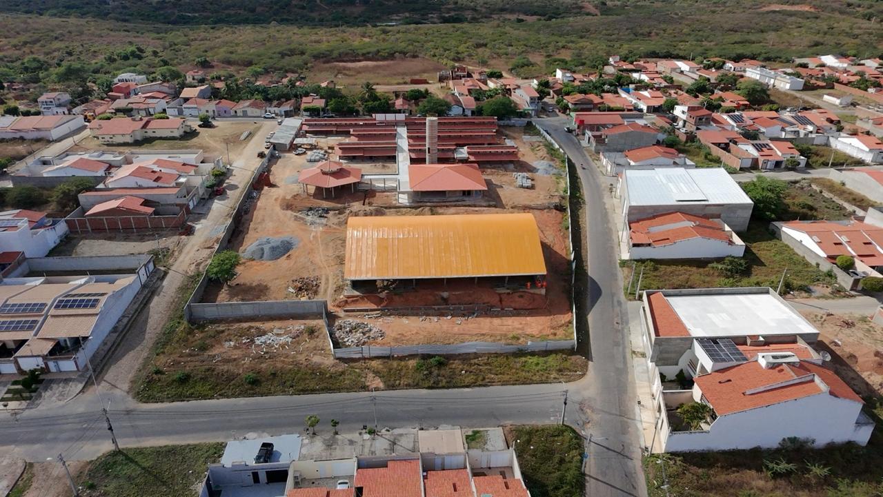 Vista aérea de uma obra em andamento, com prédios de telhado vermelho e galpão central, cercada por ruas e casas.