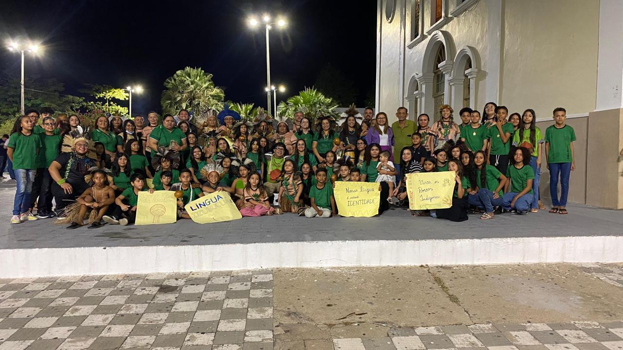 Foto de um grande grupo de indígenas, incluindo crianças e adultos, posa em frente a um prédio, segurando cartazes que valorizam a língua e a identidade indígena.