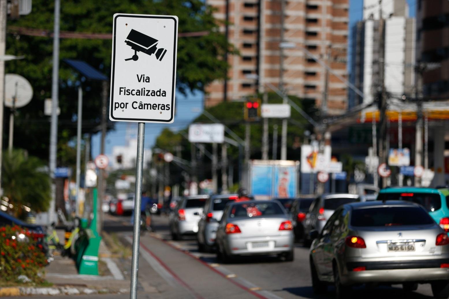 Placa de trânsito com desenho de câmera e texto ‘Via Fiscalizada por Câmeras’. Carros seguem em fila na rua movimentada. Ao fundo, prédios e semáforo no sinal vermelho.