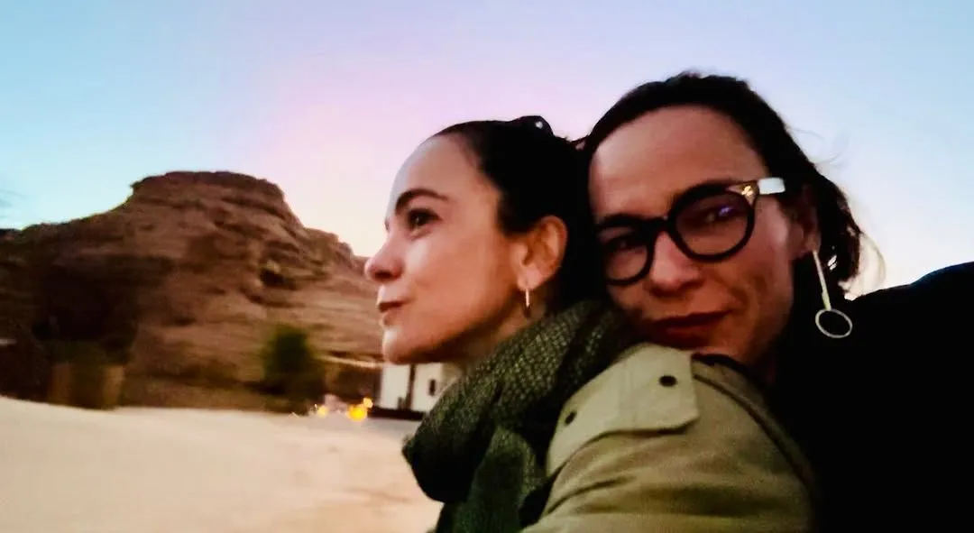 Duas mulheres abraçadas tiram uma selfie ao ar livre durante o pôr do sol. Elas estão vestidas com roupas de clima ameno, uma usando óculos e brincos longos, e a outra com cabelo preso e cachecol. Ao fundo, há uma formação rochosa e uma pequena construção, com o céu em tons de azul e rosa. A imagem transmite um momento de carinho e tranquilidade.