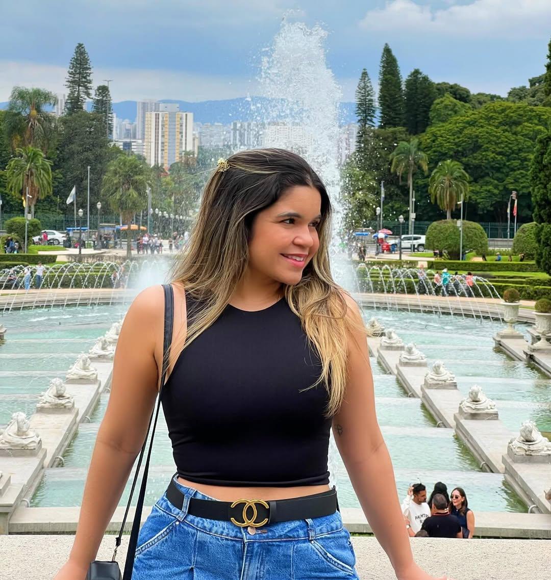 Mulher sorridente posa em frente a uma fonte no Jardim do Museu do Ipiranga, em São Paulo. Ela veste top preto, calça jeans de cintura alta com cinto preto e fivela dourada, e carrega uma bolsa de alça no ombro. Ao fundo, há fileiras de jatos d'água, esculturas de leões, árvores, palmeiras e prédios altos ao longe. O céu está nublado e há visitantes passeando no local.