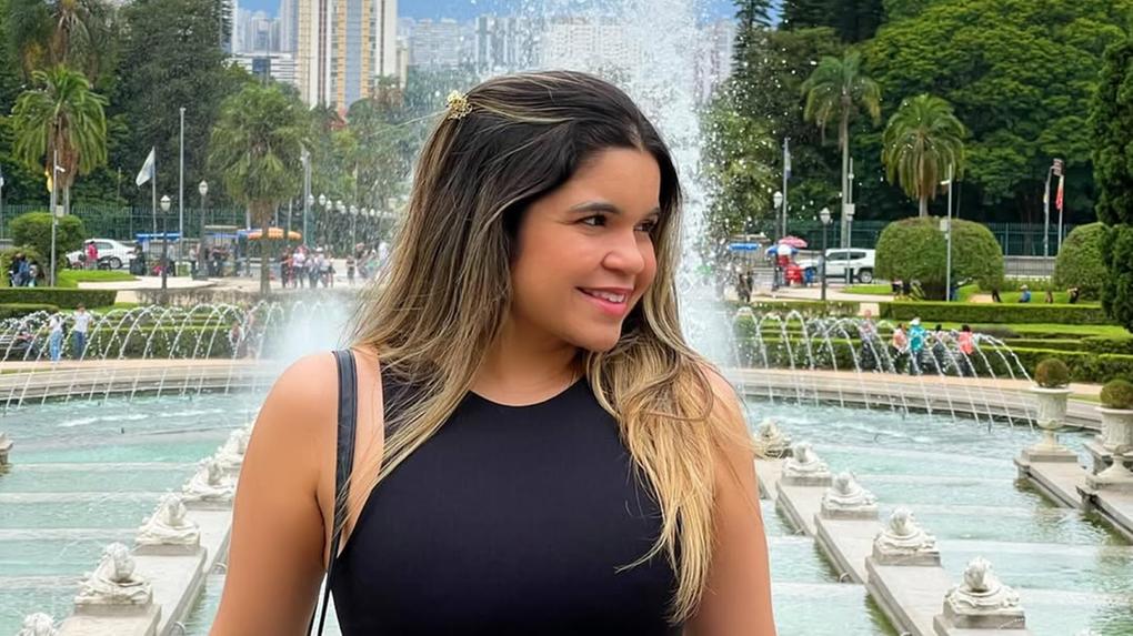 Mulher sorridente posa em frente a uma fonte no Jardim do Museu do Ipiranga, em São Paulo. Ela veste top preto, calça jeans de cintura alta com cinto preto e fivela dourada, e carrega uma bolsa de alça no ombro. Ao fundo, há fileiras de jatos d'água, esculturas de leões, árvores, palmeiras e prédios altos ao longe. O céu está nublado e há visitantes passeando no local.