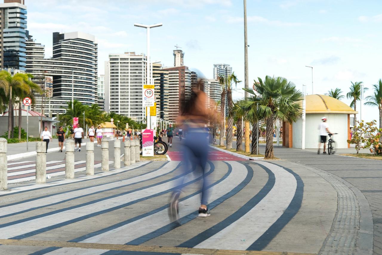 Mulher correndo na Avenida Beira Mar em Fortaleza, com prédios altos, palmeiras e movimento de pessoas ao fundo, em uma manhã ensolarada, ilustra guia de corrida para iniciantes em comemoração ao dia do Profissional de Educação Física