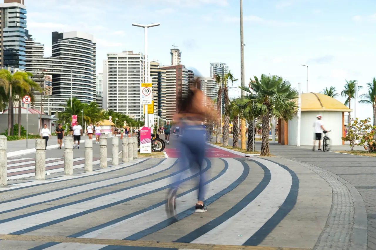 Mulher correndo na Avenida Beira Mar em Fortaleza, com prédios altos, palmeiras e movimento de pessoas ao fundo, em uma manhã ensolarada, ilustra guia de corrida para iniciantes em comemoração ao dia do Profissional de Educação Física