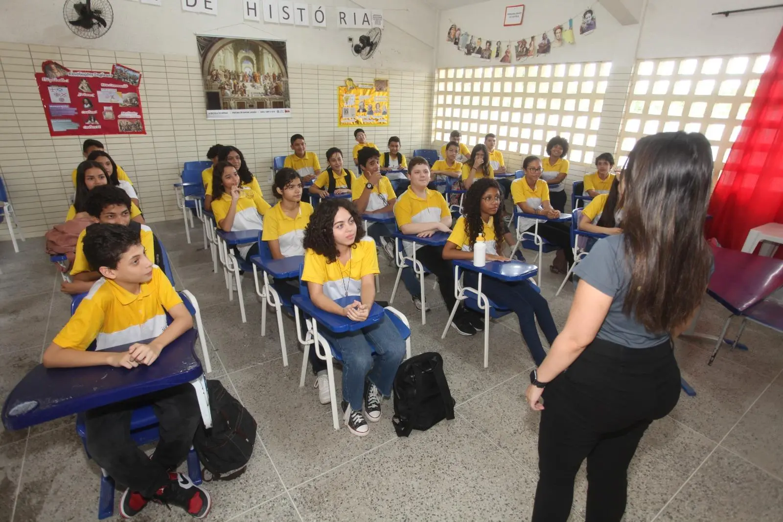 Foto que contém sala de aula com estudantes atentos à professora