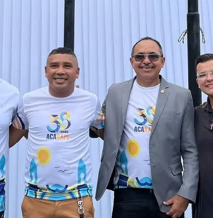 Dois homens posam sorridentes lado a lado em frente a um fundo branco com estruturas pretas. Eles vestem camisetas comemorativas brancas com estampa colorida e a inscrição 