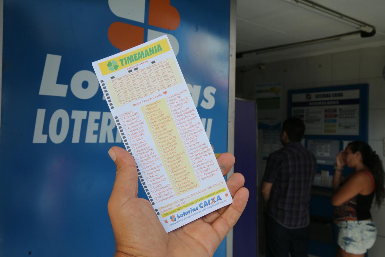 Pessoa segurando um bilhete da loteria Timemania em frente a uma casa de loteria, com fila de clientes esperando atendimento ao fundo, ilustrando loterias do dia com sorteio da caixa em 2 de setembro de 2025.