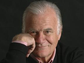 Retrato de um idoso sorridente com cabelo grisalho, usando suéter preto e camisa rosa claro, posando com a mão no rosto contra fundo preto.