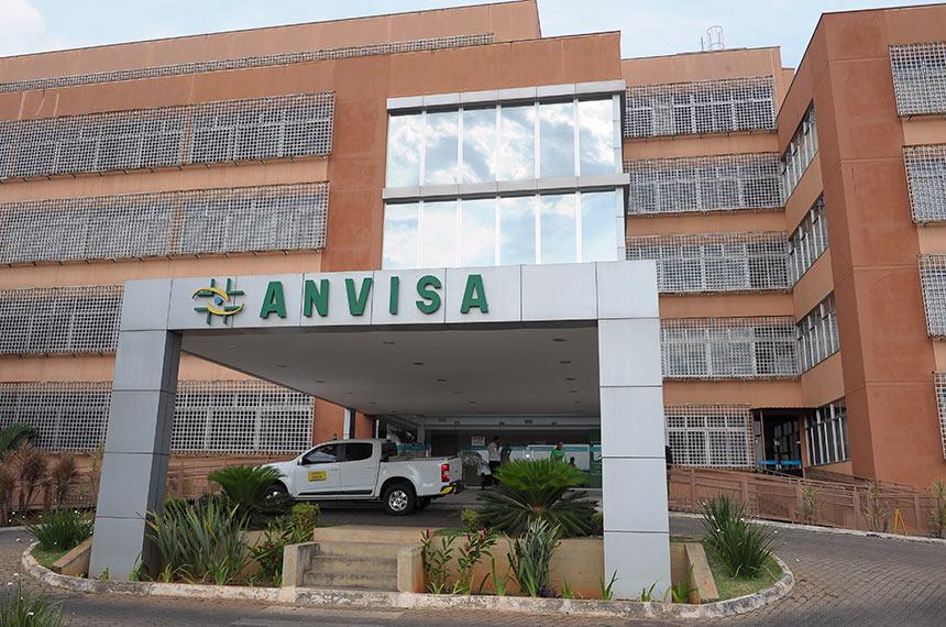 Imagem da fachada do prédio da Anvisa, agência de vigilância sanitária no Brasil, com entrada principal, estacionamento e área verde, em uma manhã ensolarada.