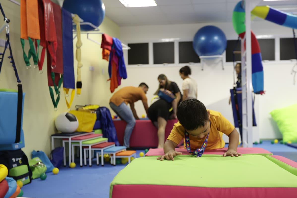 Foto de uma criança brincando em um espaço de terapia com colchonetes coloridos, enquanto adultos acompanham outras atividades ao fundo.