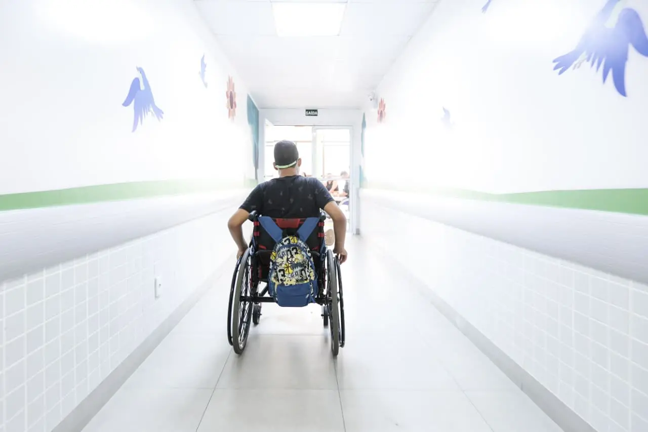 Foto de um menino em cadeira de rodas percorre um corredor branco iluminado, com paredes decoradas por desenhos coloridos.