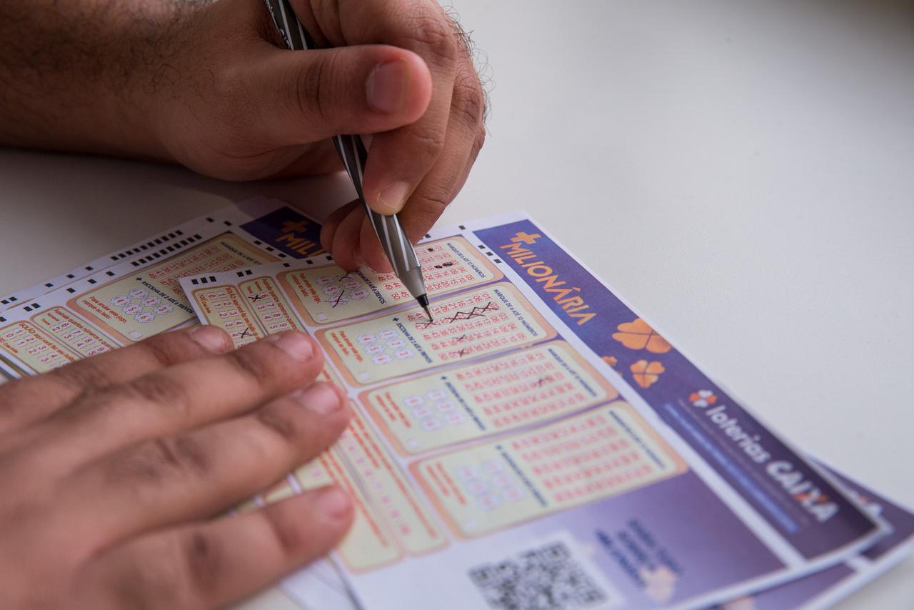 Pessoa marcando dezenas em uma cartela de jogo de loteria +Milionária, com foco na ação de escolher números e na cartela de apostas, ilustra loterias do dia com sorteio pela caixa.