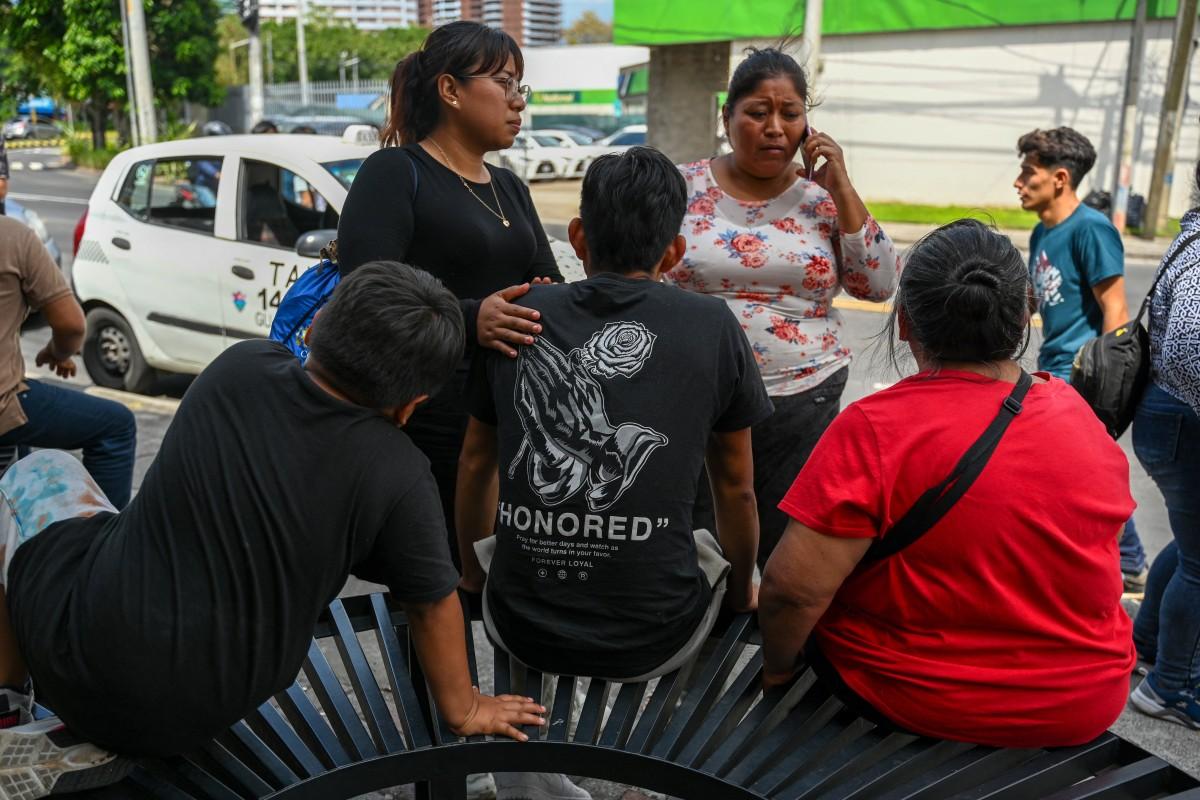 Grupo de pessoas conversando na rua, com uma mulher falando ao telefone e outras sentadas ou de pé ao redor, em um cenário urbano movimentado.