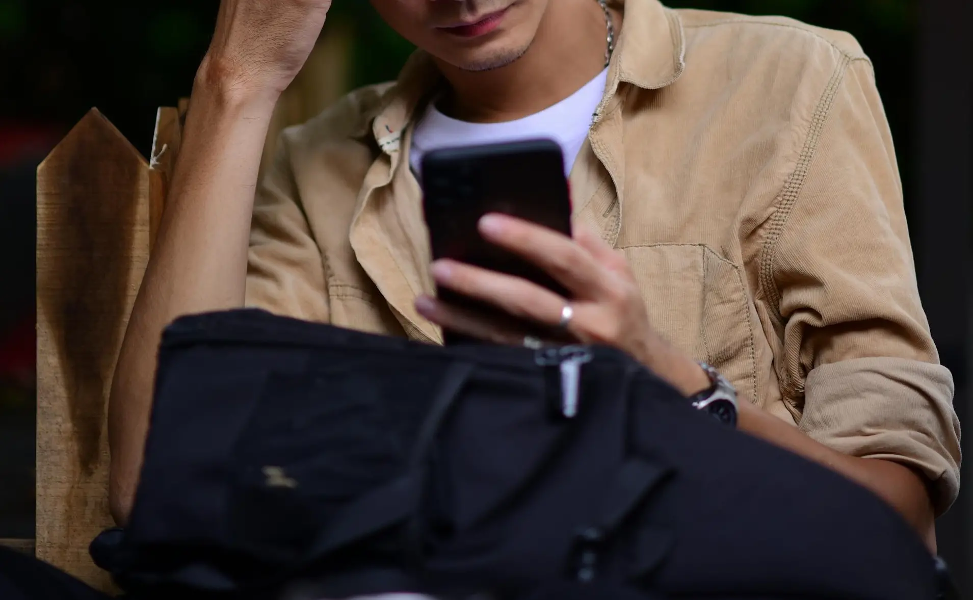 Foto de detalhe de um homem com jaqueta marrom, blusa branca, anel e relógio digitando em celular