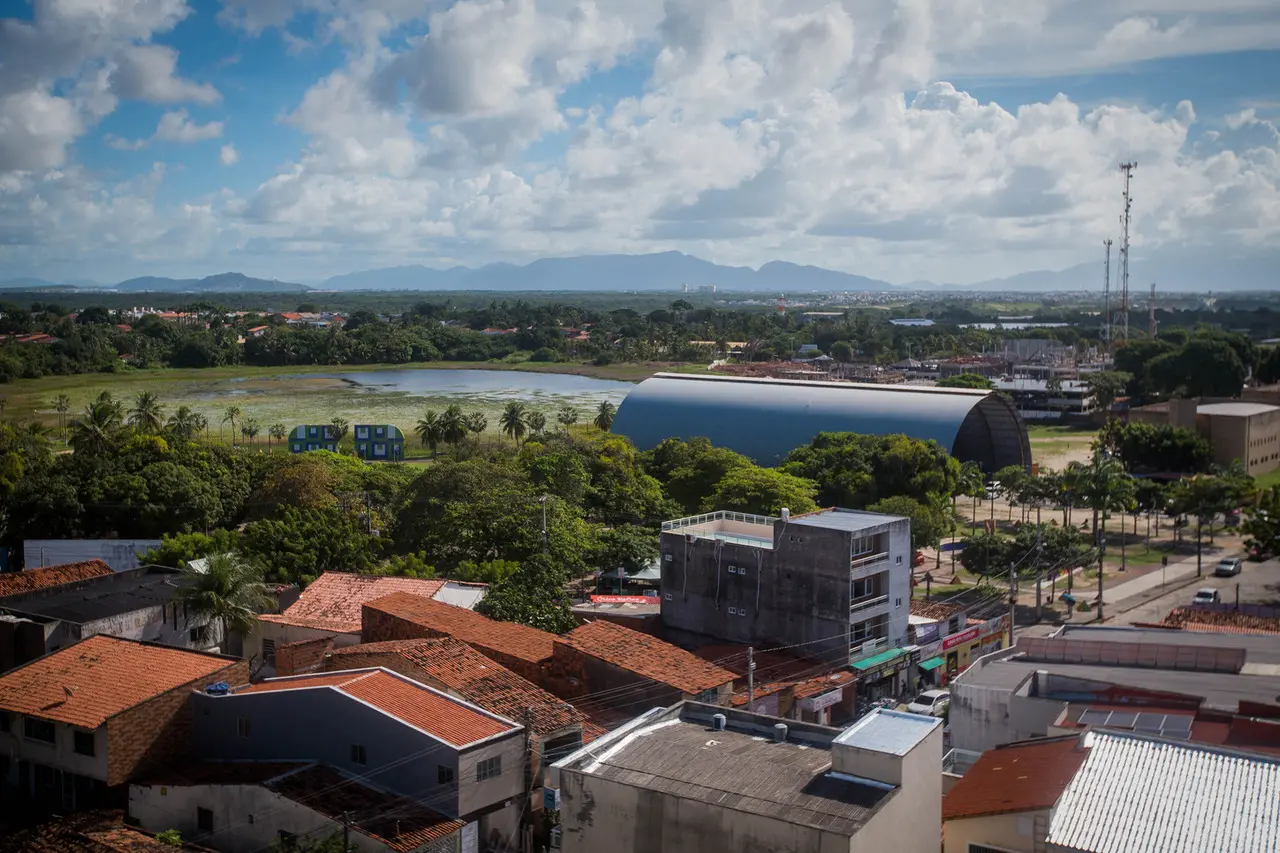 Imagem mostra vista aérea da cidade de Eusébio. Na foto, é possível ver uma lagoa e telhados de casas e empresas, além do céu azul com nuvens