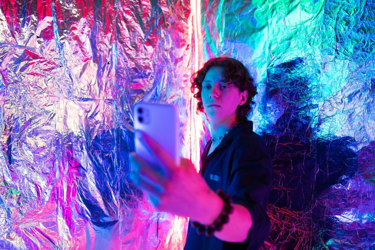 Rapaz segurança o celular na posição de quem faz selfie, ele está inserido em cenário com fundo brilhoso e colorido