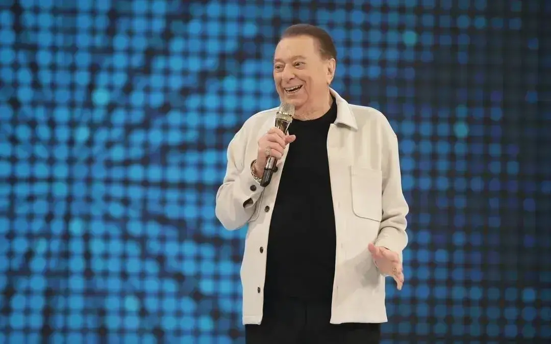 Raul Gil na frente de um painel de luzes azuis, sorrindo enquanto segura um microfone em um palco de stand-up.