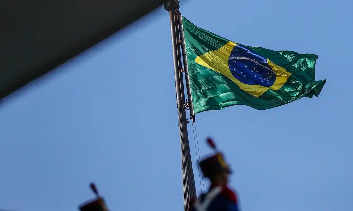Bandeira do Brasil tremulando ao vento sob céu azul, simbolizando orgulho nacional e patriotismo brasileiro.