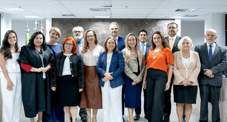 Grupo de 14 pessoas posando para foto em ambiente corporativo. Há homens e mulheres em pé, lado a lado, vestidos com roupas formais e sociais. Ao fundo, parede com revestimento geométrico e bandeiras posicionadas no canto esquerdo.