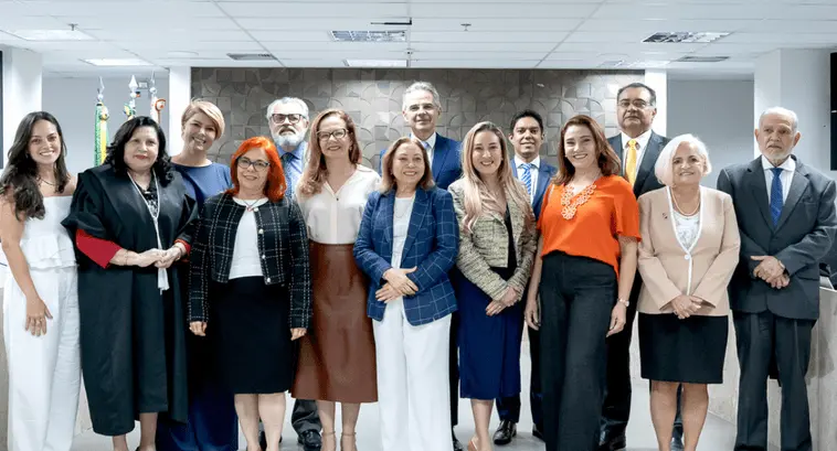 Grupo de 14 pessoas posando para foto em ambiente corporativo. Há homens e mulheres em pé, lado a lado, vestidos com roupas formais e sociais. Ao fundo, parede com revestimento geométrico e bandeiras posicionadas no canto esquerdo.