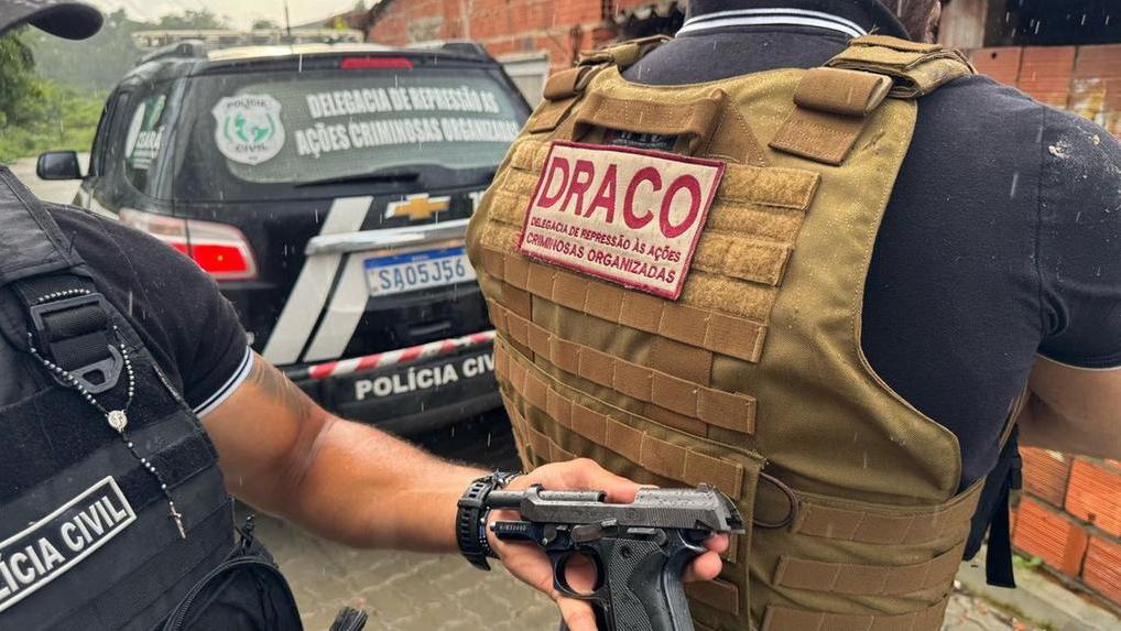 Imagem mostra policiais civis da Delegacia de Repressão às Ações Criminosas Organizadas em atuação em uma operação. A imagem mostra dois policiais civis, com coletes balísticos. Um agente mostra uma arma de fogo apreendida