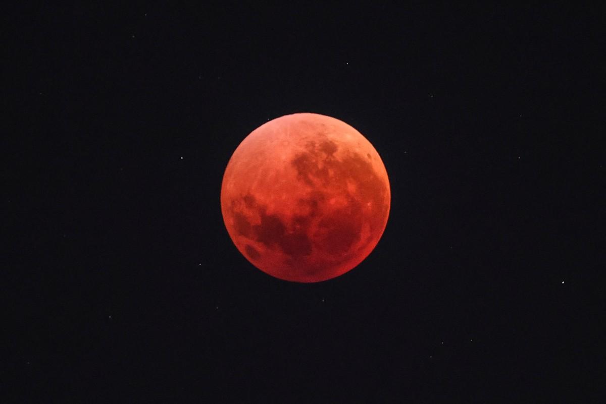 Imagem de uma superlua avermelhada, conhecida como lua de sangue, durante um eclipse lunar, com detalhes de crateras visíveis e céu escuro ao fundo.