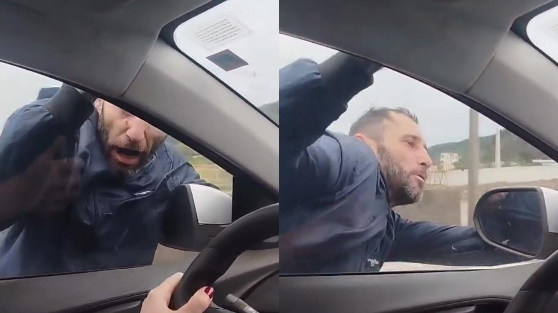 Homem com expressão de raiva tentando bater na janela do carro durante uma discussão, visto de dentro do veículo em movimento.