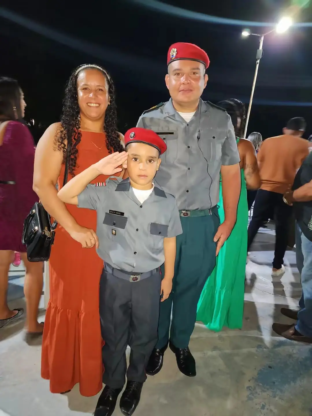 Uma fotografia que mostra um policial militar cearense, sua esposa e o filho deles, que veste o uniforme de aluno do Colégio Militar do Maranhão e faz uma saudação com a mão direita. O policial militar, à direita na foto, usa um uniforme cinza e verde-escuro e uma boina vermelha. Sua esposa está à esquerda, usando um vestido laranja. A família está posando para a foto em um evento noturno