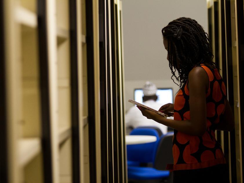 Mulher em pé entre estantes de uma biblioteca ou arquivo, segurando e lendo um livro ou caderno. Ao fundo, em uma sala com cadeiras azuis, outra pessoa está sentada em frente a um computador.