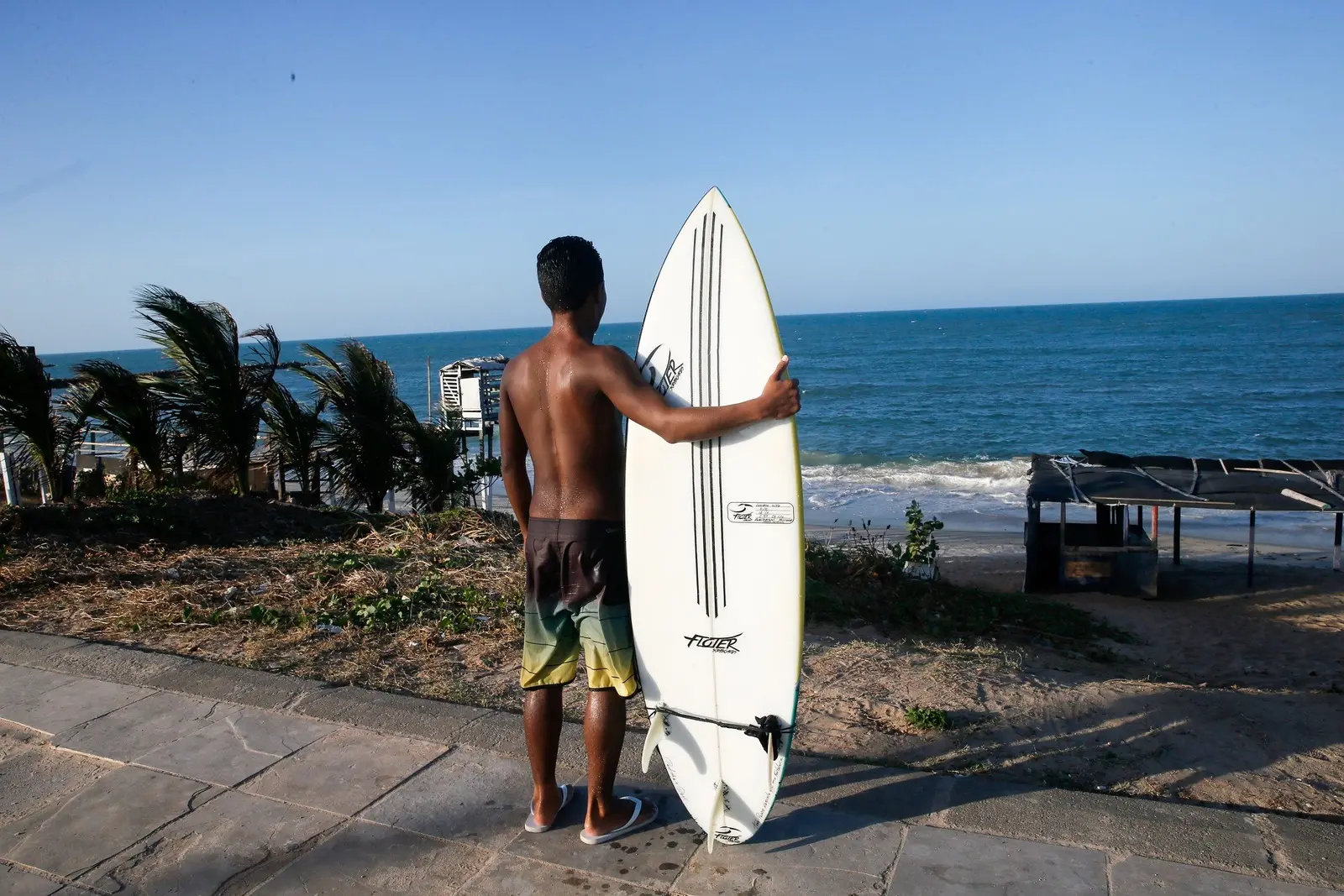 Jovem negro segurando uma prancha ao lado do corpo e olhando para o mar