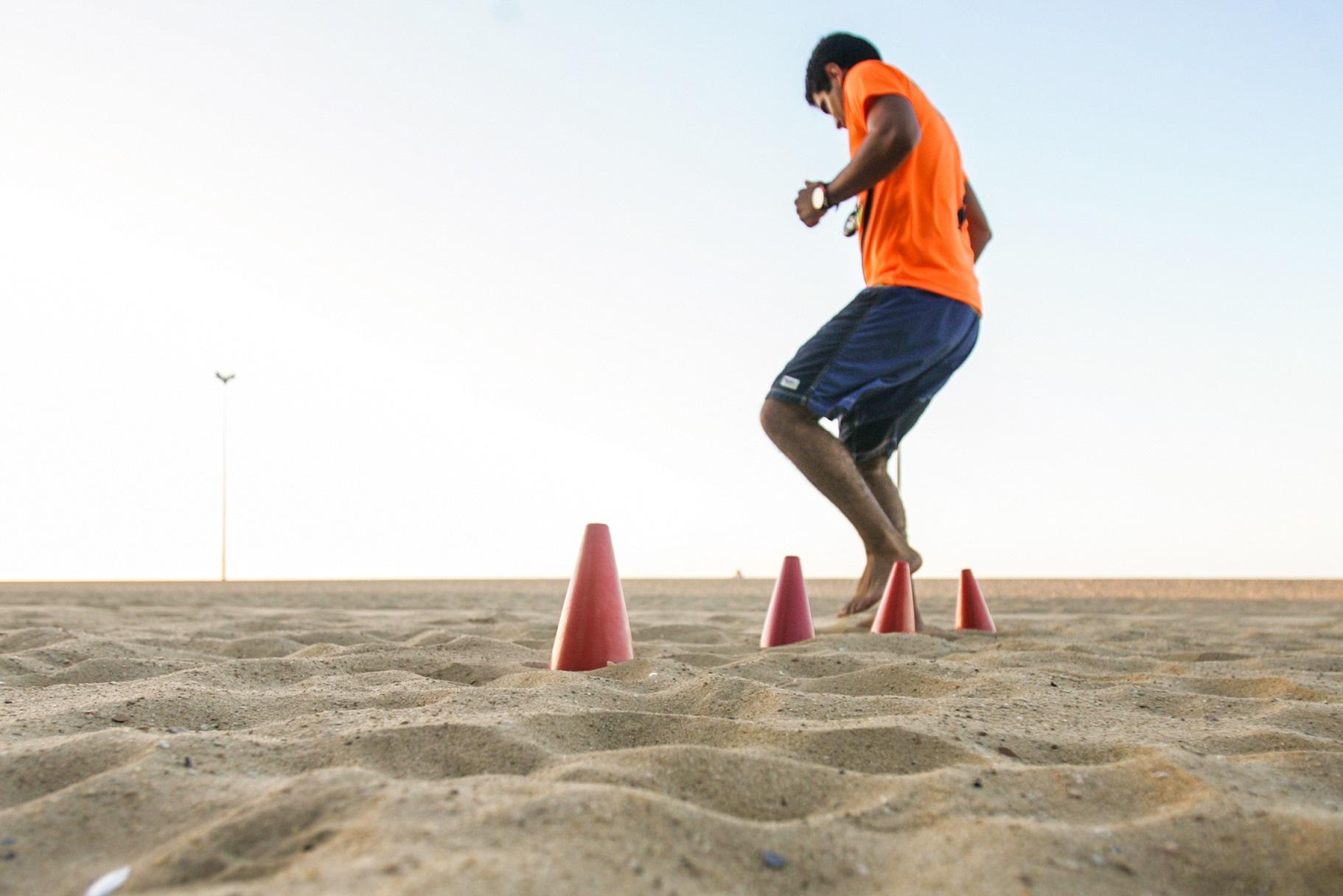 Homem de camisa laranja treinando agilidade em cones na areia