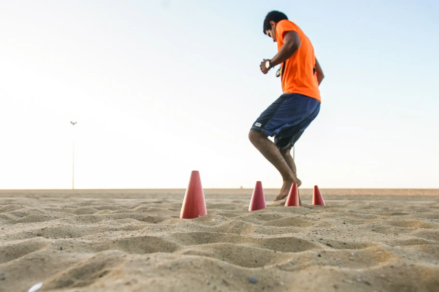 Homem de camisa laranja treinando agilidade em cones na areia