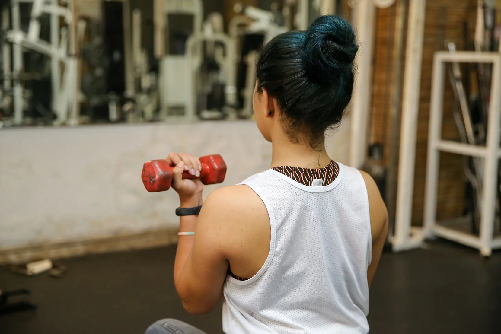 Mulher de costas de regata branca, segurando halteres vermelhos na academia