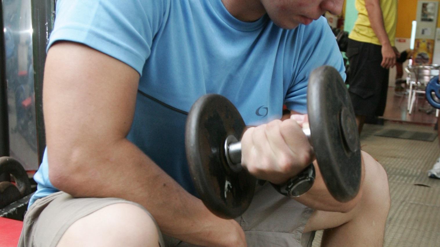 Homem jovem de camisa azul levantando um halter em um treino de braço, com concentração no exercício