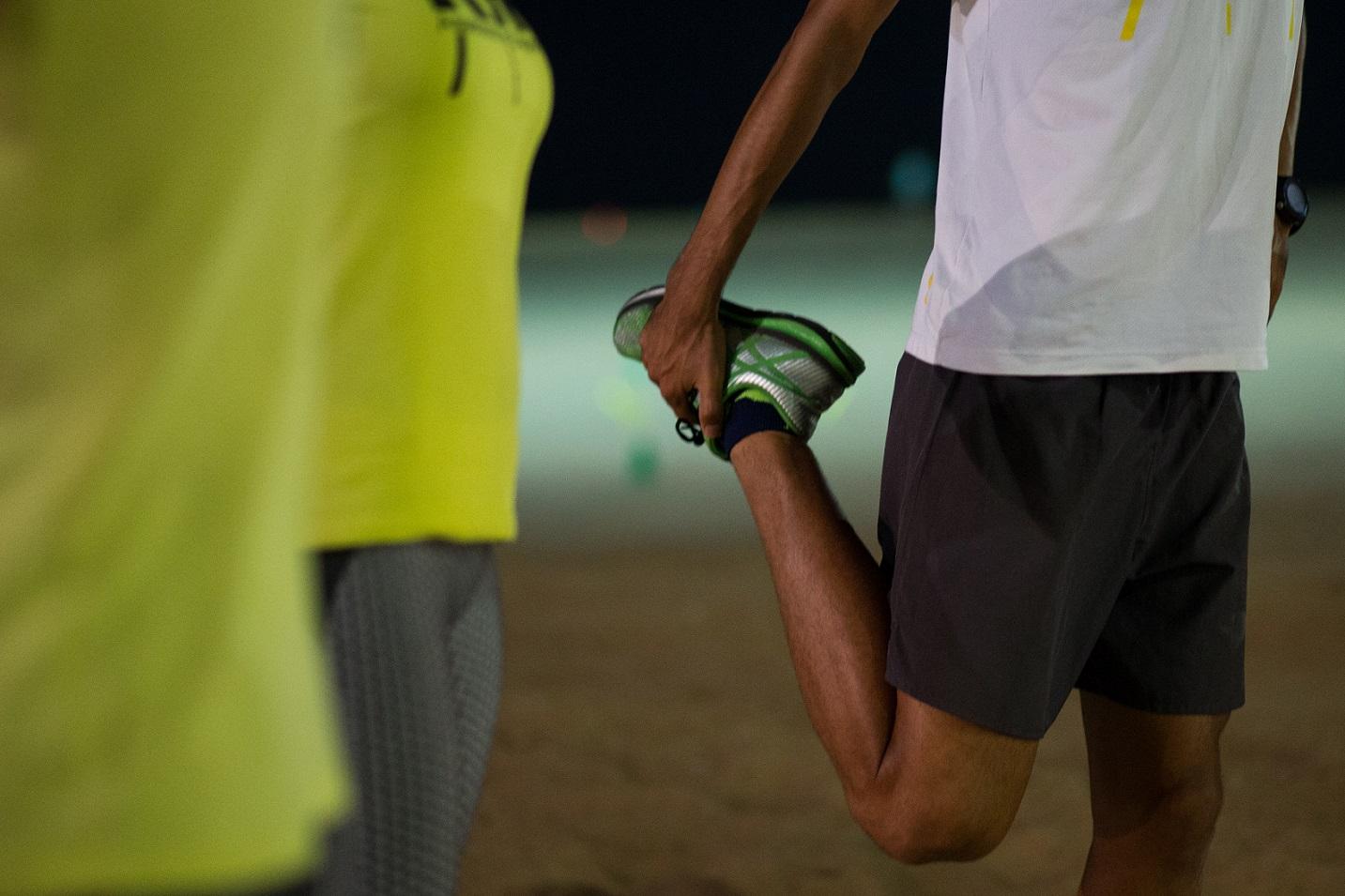 Homem alongando a perna, segurando o pé para trás. Ele usa um tênis verde e camiseta branca