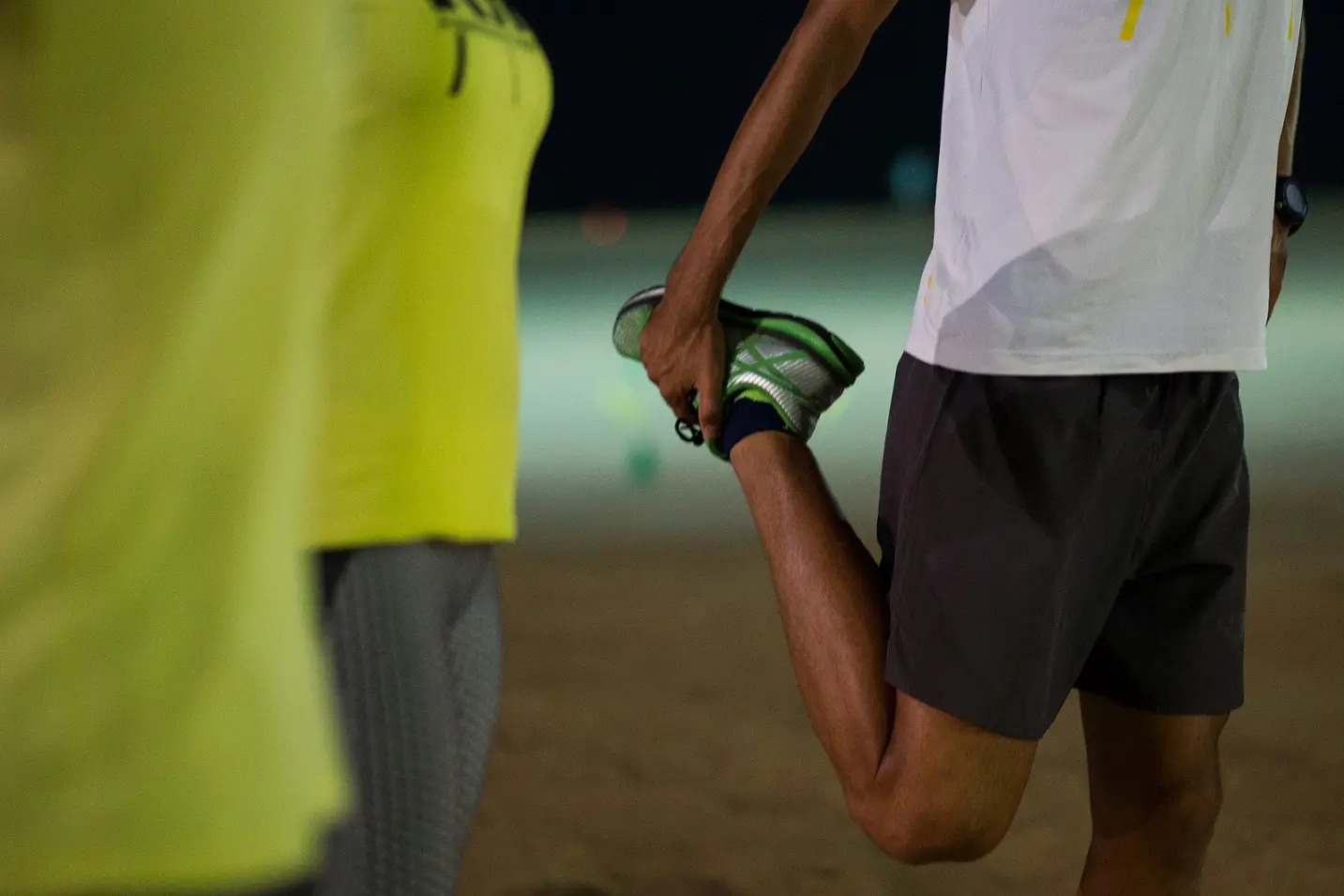Homem alongando a perna, segurando o pé para trás. Ele usa um tênis verde e camiseta branca
