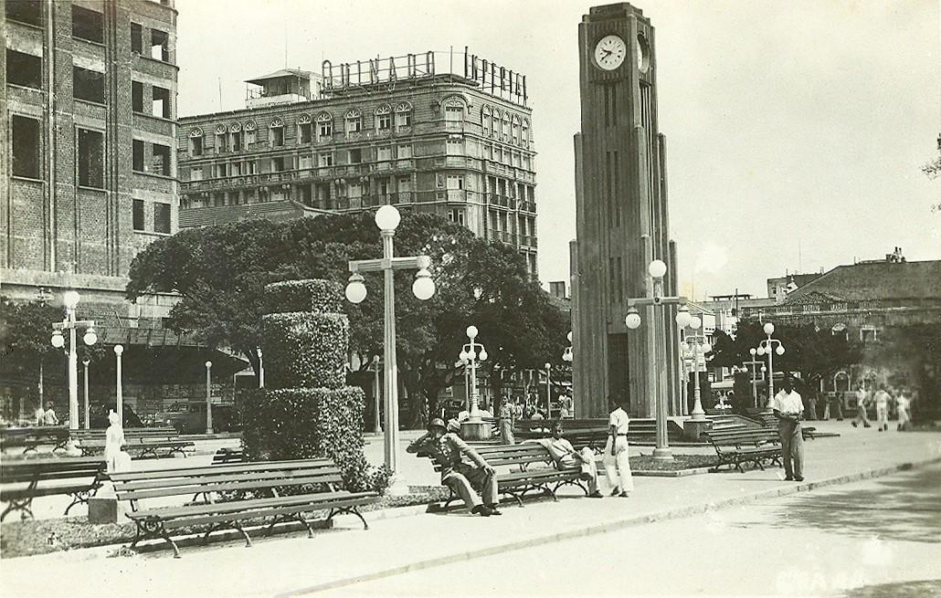 Fotografia histórica em preto e branco da Praça do Ferreira em Fortaleza. Em destaque, a Coluna da Hora, uma alta torre de relógio em estilo art déco. A praça possui bancos de madeira, postes de iluminação e árvores, com algumas pessoas em trajes de época sentadas e circulando. Ao fundo, prédios antigos compõem a paisagem urbana.
