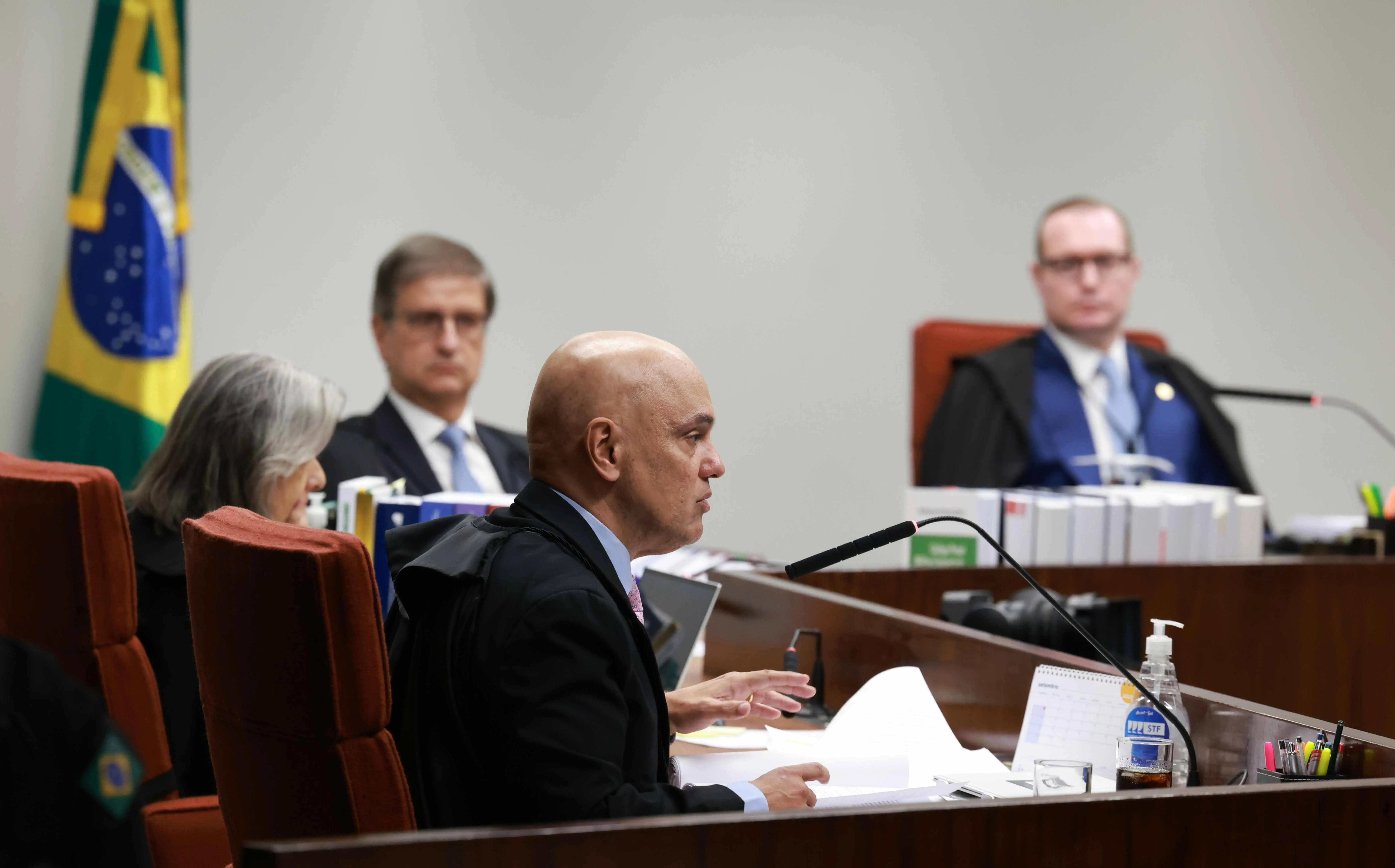 Imagem de ministros do Supremo Tribunal Federal (STF) durante sessão de julgamento. Em primeiro plano, um ministro careca fala ao microfone, segurando papéis sobre a mesa. Trata-se de Alexandre de Moraes. Ao fundo, outros ministros estão sentados, com livros, processos e materiais organizados sobre as bancadas. No canto esquerdo, parte da bandeira do Brasil é visível. Sobre a mesa da frente há um frasco de álcool em gel e canetas em um suporte.