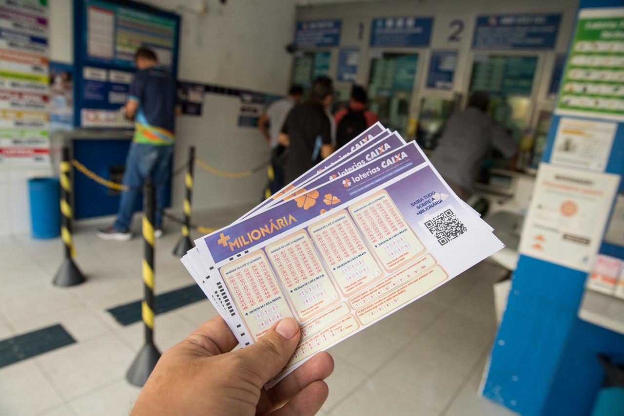 Imagem mostra pessoa segurando bilhetes de loteria +Milionária na fila de uma lotérica, ambiente com várias pessoas comprando apostas de loteria.