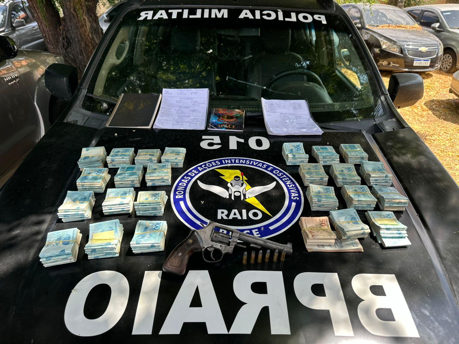 Apreensão da polícia militar em cima de parte dianteira de carro preto do Batalhão do Raio: quantidade de dinheiro, arma de fogo e cadernos de materiais ilegais.