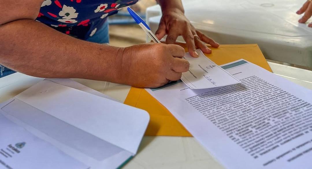 Pessoa assina um documento oficial sobre uma mesa branca. Ao lado, há um envelope amarelo e outros papéis impressos com o brasão da Prefeitura de Fortaleza, relacionados a processos de regularização fundiária.
