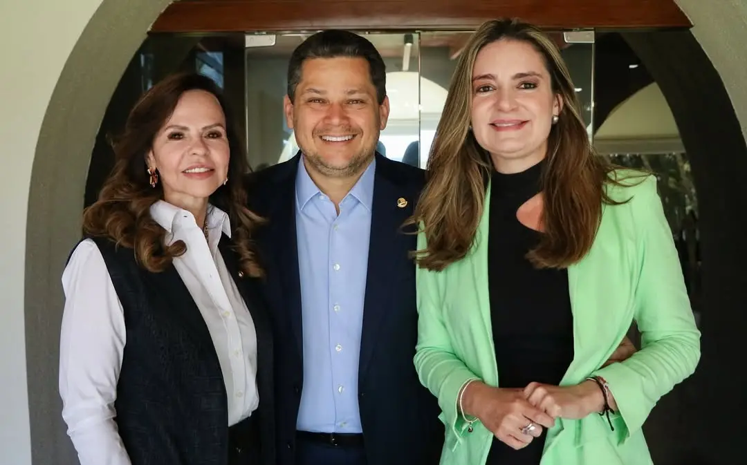 Duas mulheres e um homem posam sorridentes para foto em ambiente interno. A mulher à esquerda usa camisa branca e colete preto, o homem ao centro veste terno azul e camisa clara, e a mulher à direita está de blazer verde sobre blusa preta