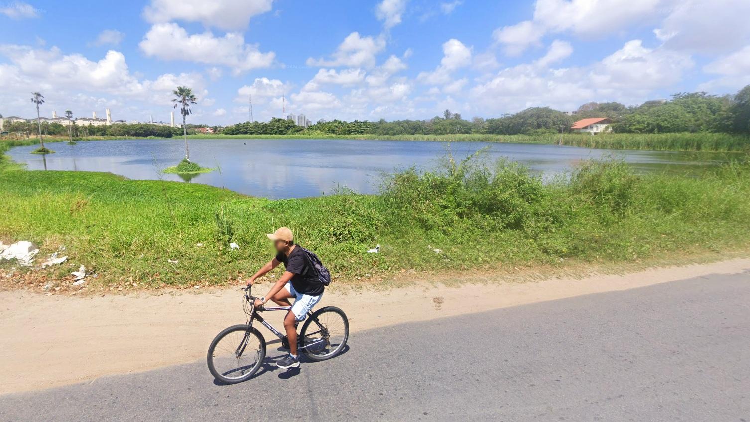 Imagem mostra homem andando de bicicleta em uma rua à frente de uma lagoa em Fortaleza. Vegetação cerca a margem da lagoa.