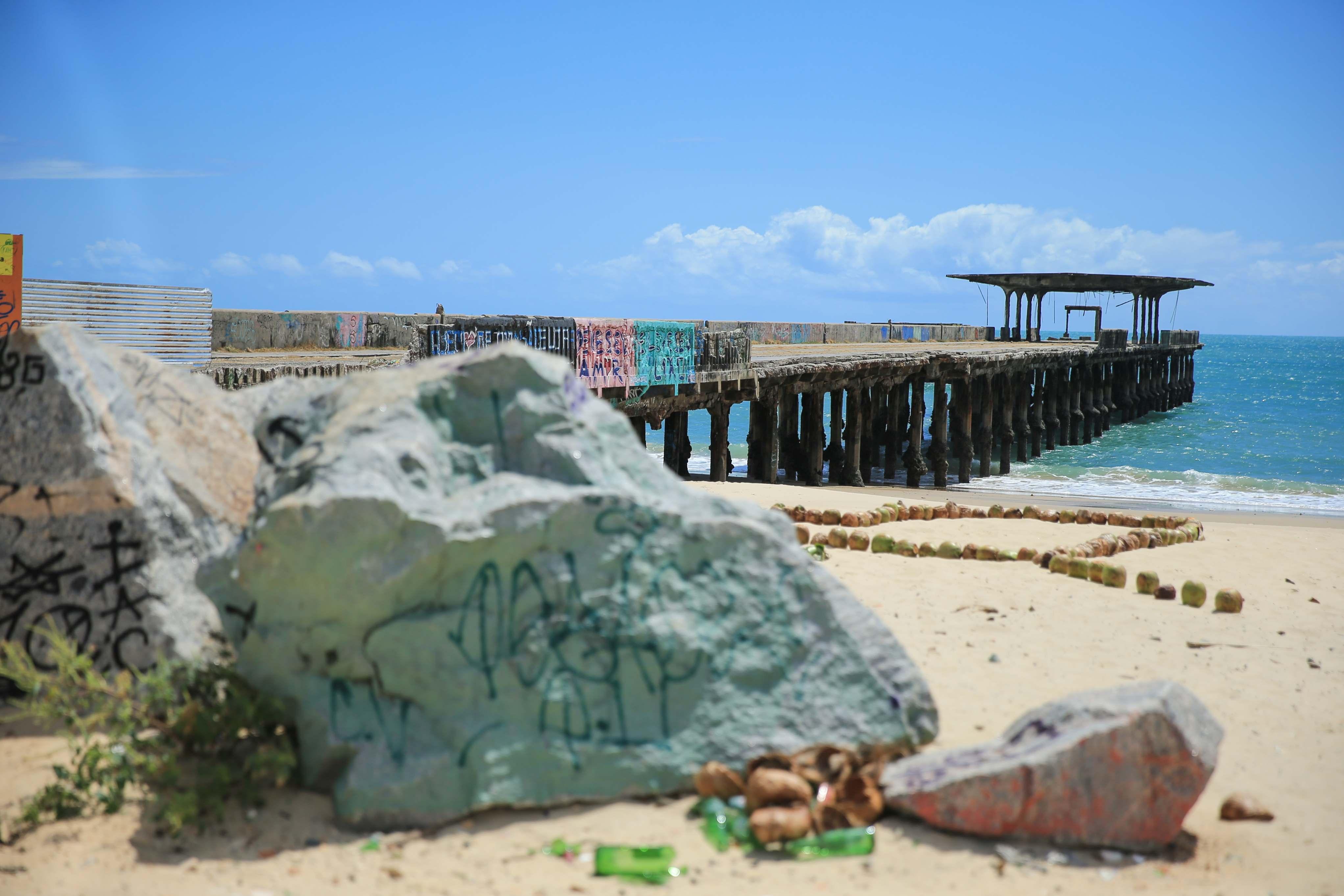 Vista do antigo cais de Iracema em Fortaleza, Ceará. Em primeiro plano, pedras grandes com grafites e alguns detritos na areia da praia. Ao fundo, o cais de madeira e concreto se estende sobre o mar azul-turquesa, com um gazebo em ruínas na ponta e parte de sua estrutura coberta por grafites coloridos. O céu é predominantemente azul com algumas nuvens brancas.