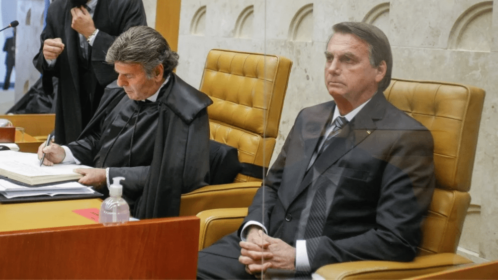 Dois homens estão sentados em cadeiras de couro na cor bege, em um ambiente formal de tribunal. À esquerda, Fux, ministro do Supremo Tribunal Federal (STF), veste toga preta e escreve em um livro. À direita, um homem de terno escuro e gravata listrada permanece sentado com expressão séria. Trata-se de Jair Bolsonaro. Entre eles, há uma divisória de acrílico transparente. Em primeiro plano, sobre a mesa, há um frasco de álcool em gel.