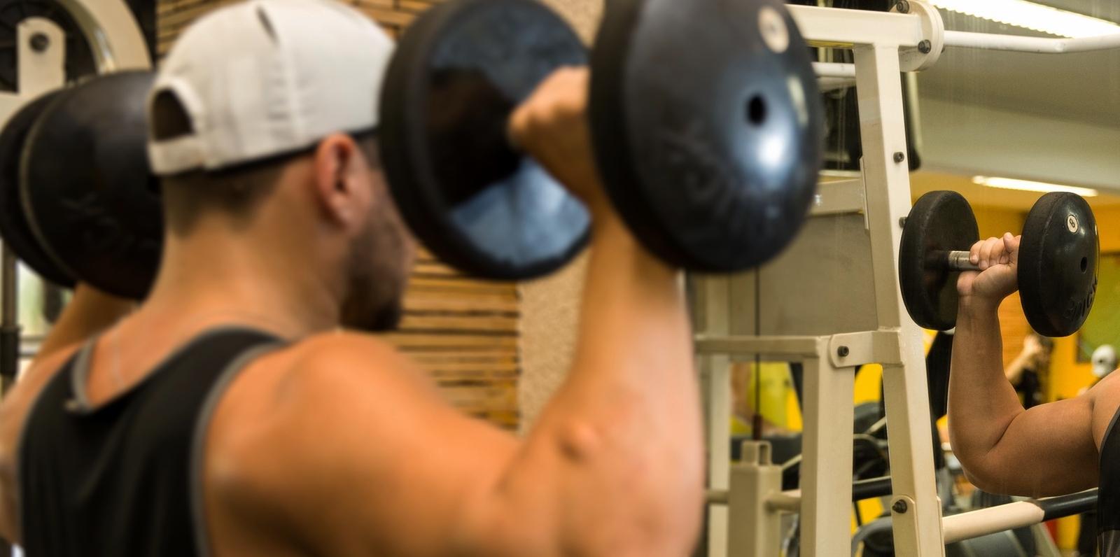 Homem musculoso de boné branco e regata preta faz exercício para ombros em frente a espelho de academia