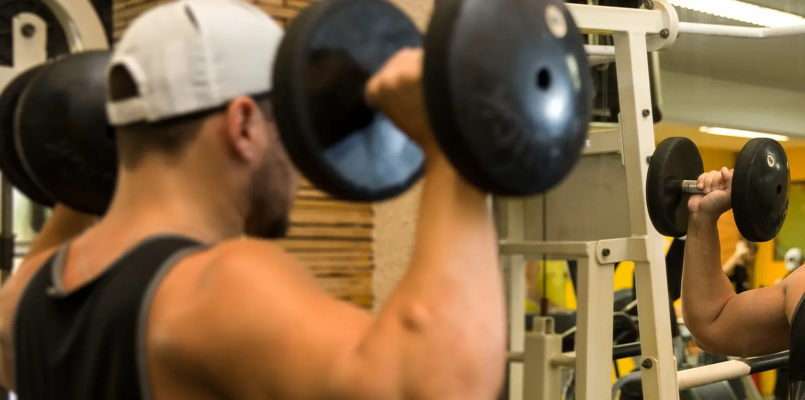 Homem musculoso de boné branco e regata preta faz exercício para ombros em frente a espelho de academia